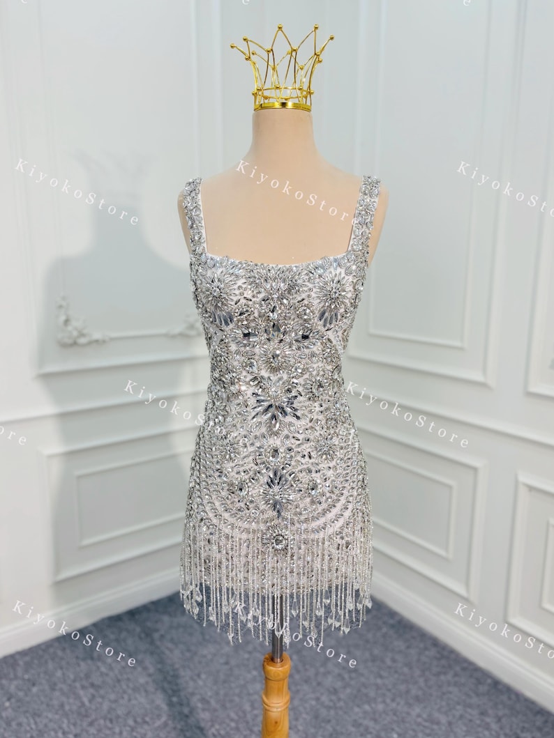 K&ouml;nnte beinhalten: Ein silbernes, perlenbesetztes Minikleid mit quadratischem Ausschnitt und Fransen. Das Kleid ist mit glitzernden Verzierungen versehen und wird auf einer Schaufensterpuppe mit goldener Krone pr&auml;sentiert. Das Kleid ist etwa 76 cm lang.