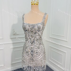 K&ouml;nnte beinhalten: Ein kurzes, wei&szlig;es Kleid mit aufwendigen silbernen Perlen und Fransen. Das Kleid hat einen quadratischen Ausschnitt und wird auf einer Schaufensterpuppe mit einer goldenen Krone pr&auml;sentiert. Ein glamour&ouml;ses Outfit f&uuml;r besondere Anl&auml;sse.