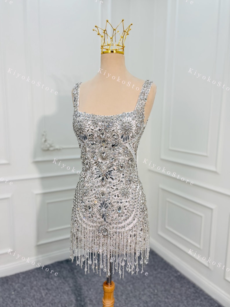 K&ouml;nnte beinhalten: Ein kurzes, wei&szlig;es Kleid mit aufwendigen silbernen Perlen und Fransen. Das Kleid hat einen quadratischen Ausschnitt und wird auf einer Schaufensterpuppe mit einer goldenen Krone pr&auml;sentiert. Ein glamour&ouml;ses Outfit f&uuml;r besondere Anl&auml;sse.