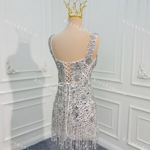 K&ouml;nnte beinhalten: Ein silbernes, perlenbesetztes Minikleid mit Schn&uuml;rr&uuml;cken und Fransen. Das Kleid wird auf einer Schaufensterpuppe mit einer goldenen Krone pr&auml;sentiert. Das Kleid ist mit glitzernden Pailletten und Perlen verziert.