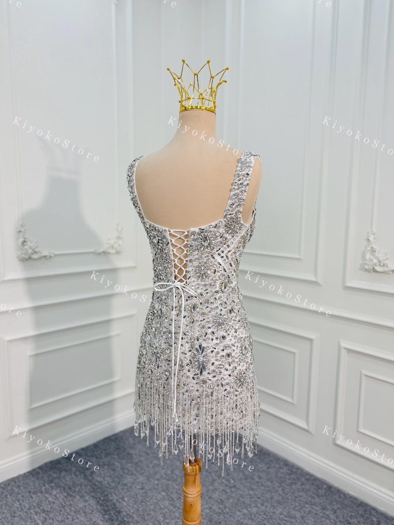 K&ouml;nnte beinhalten: Ein silbernes, perlenbesetztes Minikleid mit Schn&uuml;rr&uuml;cken und Fransen. Das Kleid wird auf einer Schaufensterpuppe mit einer goldenen Krone pr&auml;sentiert. Das Kleid ist mit glitzernden Pailletten und Perlen verziert.