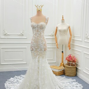 Pu&ograve; includere: Un abito da sposa a sirena bianco lungo fino a terra con corpetto trasparente, applicazioni in pizzo floreale, scollo a cuore, spalline in pizzo spesso e gonna in tulle svasata con un lungo strascico, esposto su un manichino con corona.