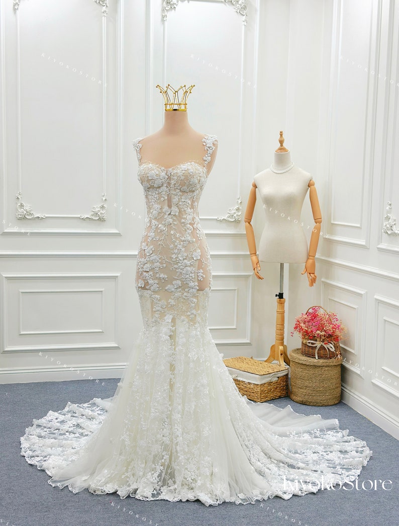 Pu&ograve; includere: Un abito da sposa a sirena bianco lungo fino a terra con corpetto trasparente, applicazioni in pizzo floreale, scollo a cuore, spalline in pizzo spesso e gonna in tulle svasata con un lungo strascico, esposto su un manichino con corona.