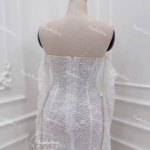 May include: A white wedding dress with long sleeves and a fitted bodice. The dress is covered in delicate beading and lace.