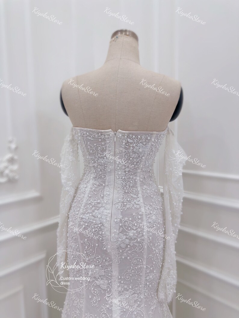 May include: A white wedding dress with long sleeves and a fitted bodice. The dress is covered in delicate beading and lace.