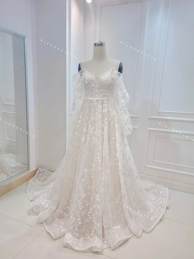 May include: A white off-the-shoulder wedding dress with a sweetheart neckline and a tulle skirt. The dress is covered in small white stars.