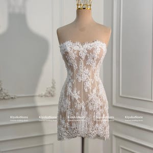 May include: A strapless mini dress featuring a nude-colored base with intricate white floral lace overlays, delicate sequin embellishments, and a fringe hemline. The garment is displayed on a mannequin wearing a gold-toned wire crown.