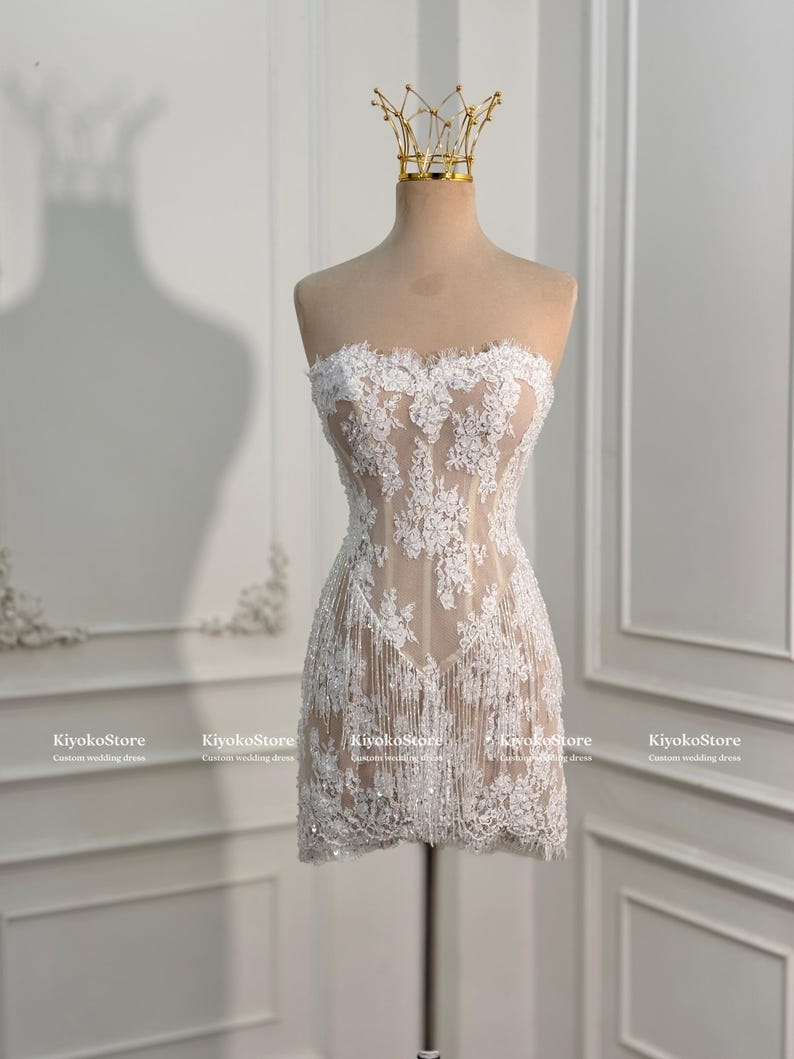 May include: A strapless mini dress featuring a nude-colored base with intricate white floral lace overlays, delicate sequin embellishments, and a fringe hemline. The garment is displayed on a mannequin wearing a gold-toned wire crown.