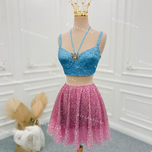 Puede incluir: Un conjunto formal de dos piezas sobre un maniquí. La parte superior es un bustier azul claro con lentejuelas, con un collar a juego y un colgante de tigre. La falda es una minifalda rosa con lentejuelas. Una corona dorada está sobre la cabeza del maniquí.