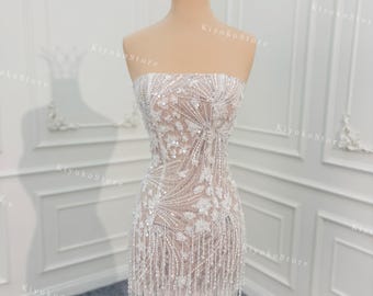 May include: A strapless, white beaded mini dress with floral and linear patterns. The dress features a fringe hem and is displayed on a mannequin. A gold crown sits atop the mannequin's head. The background is white.