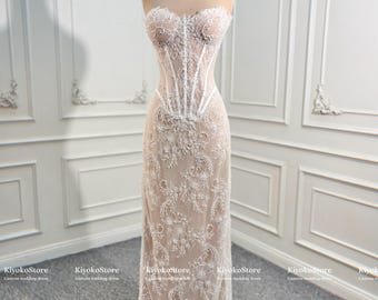 May include: A strapless floor-length gown featuring a nude-colored base with intricate white lace embroidery and a structured corset-style bodice. The dress is displayed on a mannequin wearing a gold-toned crown, with KiyokoStore text visible.