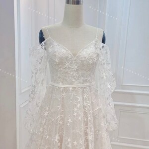 May include: A white wedding dress with a star-shaped pattern and off-the-shoulder straps. The dress has a fitted bodice and a flowing skirt.