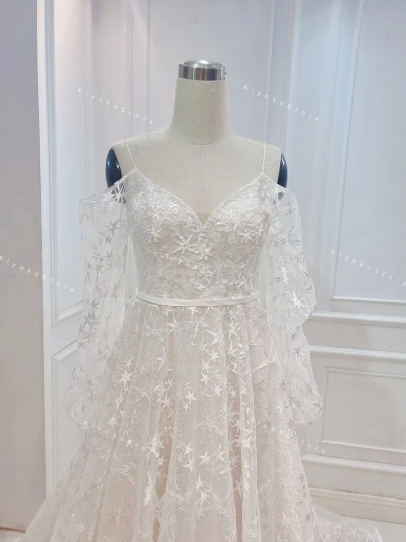 May include: A white wedding dress with a star-shaped pattern and off-the-shoulder straps. The dress has a fitted bodice and a flowing skirt.