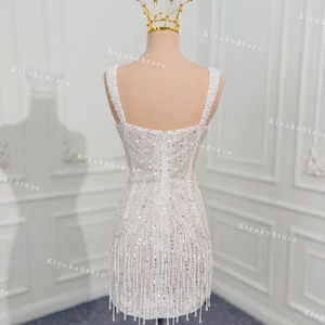 May include: A white, beaded mini dress with a square neckline and fringe detail. The dress is displayed on a mannequin with a gold crown. The dress has a fitted bodice and a short skirt, perfect for a special occasion.