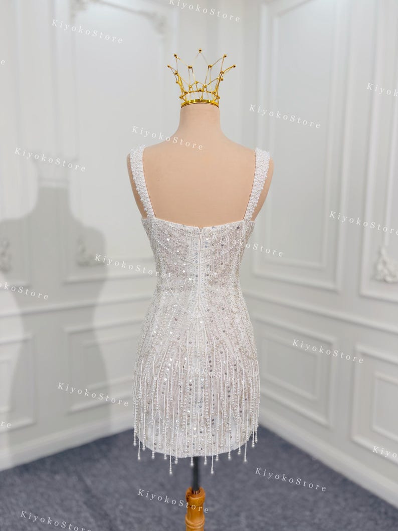 May include: A white, beaded mini dress with a square neckline and fringe detail. The dress is displayed on a mannequin with a gold crown. The dress has a fitted bodice and a short skirt, perfect for a special occasion.