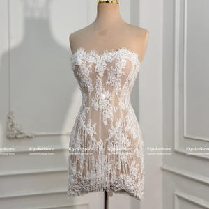 May include: A strapless mini dress featuring a nude-colored base with white floral lace overlays, intricate embroidery, and a fringe hemline. The dress is displayed on a mannequin wearing a gold-colored wire crown. Text reads KiyokoStore.