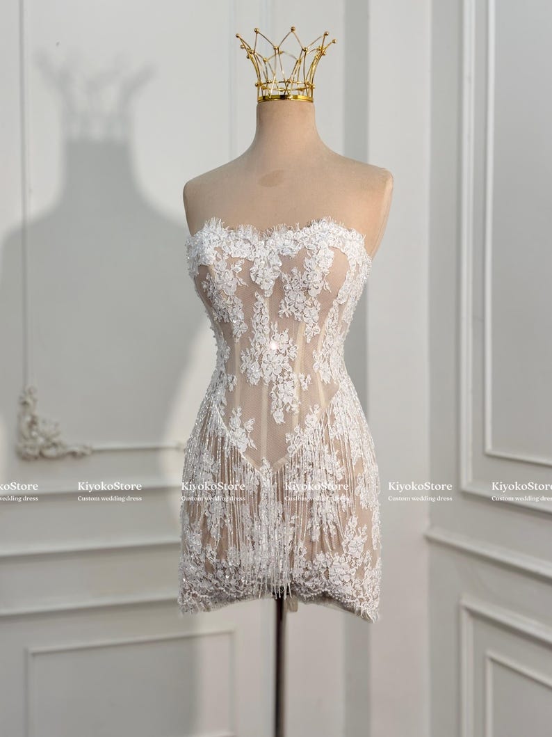 May include: A strapless mini dress featuring a nude-colored base with white floral lace overlays, intricate embroidery, and a fringe hemline. The dress is displayed on a mannequin wearing a gold-colored wire crown. Text reads KiyokoStore.