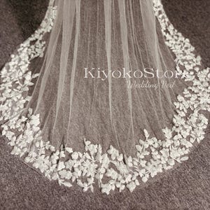 May include: A white wedding veil with a delicate floral lace trim. The veil is made of soft tulle and has a scalloped edge. The lace trim features small white flowers and leaves.