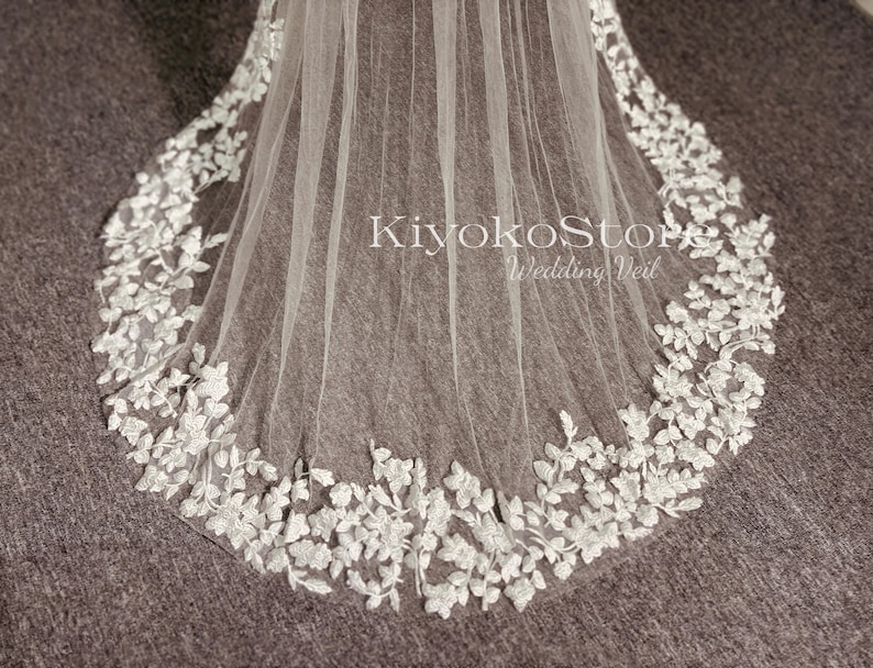 May include: A white wedding veil with a delicate floral lace trim. The veil is made of soft tulle and has a scalloped edge. The lace trim features small white flowers and leaves.