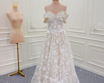 May include: A white off-the-shoulder wedding dress with floral embellishments. The gown is displayed on a mannequin, showcasing its intricate details and flowing skirt. A gold crown sits on a nearby mannequin.
