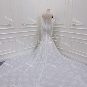 May include: A white wedding dress with long sleeves and a fitted bodice. The dress has a lace overlay with a floral pattern and a long train.