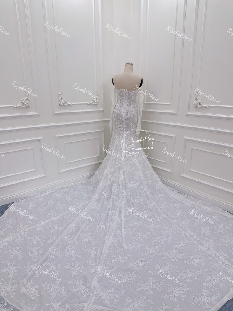 May include: A white wedding dress with long sleeves and a fitted bodice. The dress has a lace overlay with a floral pattern and a long train.