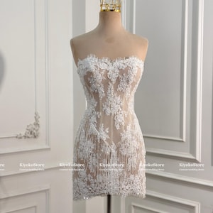 May include: A strapless mini dress featuring a nude-colored base with intricate white floral lace overlays and delicate fringe detailing along the hem. The garment is displayed on a mannequin topped with a gold-colored wire crown accessory.