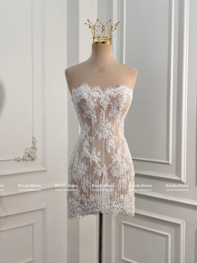 May include: A strapless mini dress featuring a nude-colored base with intricate white floral lace overlays and delicate fringe detailing along the hem. The garment is displayed on a mannequin topped with a gold-colored wire crown accessory.