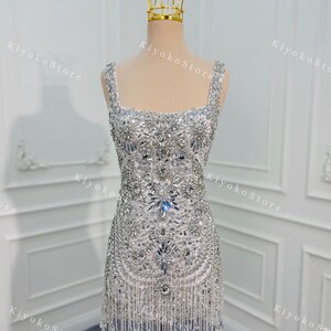 K&ouml;nnte beinhalten: Ein silbernes, perlenbesetztes Minikleid mit quadratischem Ausschnitt und Fransen. Das Kleid wird auf einer Schaufensterpuppe mit einer goldenen Krone pr&auml;sentiert. Das Kleid ist stark mit glitzernden Kristallen und Perlen verziert.