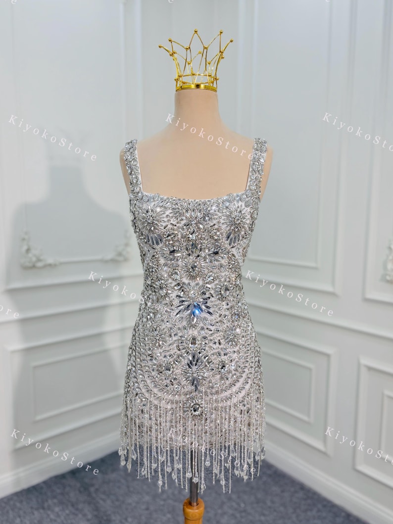 K&ouml;nnte beinhalten: Ein silbernes, perlenbesetztes Minikleid mit quadratischem Ausschnitt und Fransen. Das Kleid wird auf einer Schaufensterpuppe mit einer goldenen Krone pr&auml;sentiert. Das Kleid ist stark mit glitzernden Kristallen und Perlen verziert.