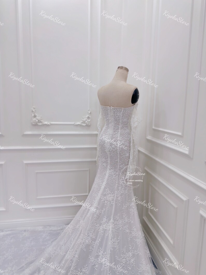 May include: A white wedding dress with a fitted bodice and a flowing skirt. The dress is covered in delicate lace and beading. The dress has a sweetheart neckline and a long train.