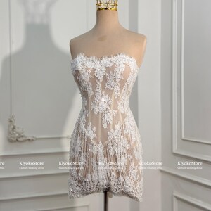 May include: A strapless mini dress featuring a nude-colored base layer with intricate white floral lace overlays and delicate fringe detailing along the hem. The garment is displayed on a mannequin topped with a gold-toned wire crown accessory.