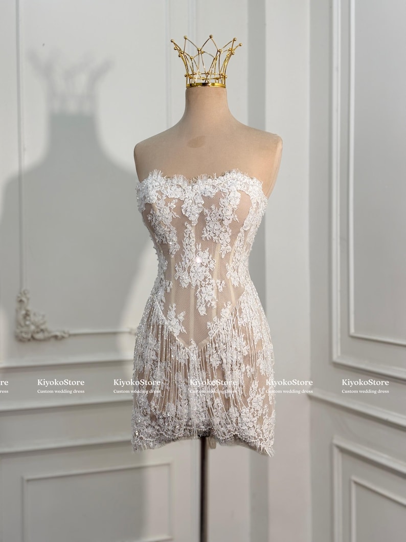 May include: A strapless mini dress featuring a nude-colored base layer with intricate white floral lace overlays and delicate fringe detailing along the hem. The garment is displayed on a mannequin topped with a gold-toned wire crown accessory.