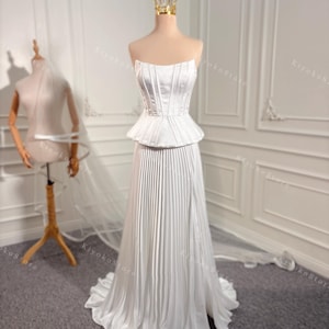 May include: A white strapless wedding dress with a fitted bodice and a pleated skirt. The skirt has a high slit on the left side. The dress is on a mannequin in a white room.