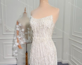 May include: A white strapless mini dress with a beaded bodice and a fringe hem. The dress is on a mannequin and has a crown on top of its head.
