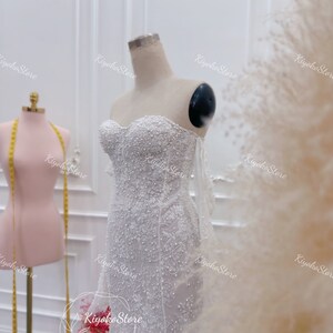 May include: A white beaded wedding dress with a sweetheart neckline and a fitted silhouette. The dress has a sheer overlay with delicate beading and a train.