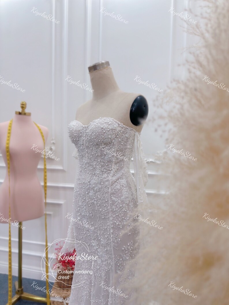 May include: A white beaded wedding dress with a sweetheart neckline and a fitted silhouette. The dress has a sheer overlay with delicate beading and a train.
