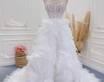 May include: A strapless white wedding dress with a lace bodice and a pearl-beaded neckline. The skirt features layers of ruffled fabric, creating a voluminous effect. The dress is displayed on a mannequin with a gold stand. The text "Kiyoko Store Wedding dress" is visible.