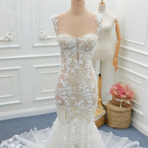 Pu&ograve; includere: Un abito a sirena lungo fino a terra con base color carne, applicazioni in pizzo floreale bianco, scollo a cuore e spalline sottili. Il corpetto aderente si allarga in un ampio strascico in tulle trasparente sul fondo.