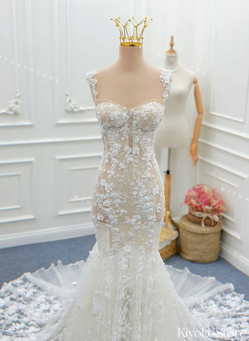 Pu&ograve; includere: Un abito a sirena lungo fino a terra con base color carne, applicazioni in pizzo floreale bianco, scollo a cuore e spalline sottili. Il corpetto aderente si allarga in un ampio strascico in tulle trasparente sul fondo.