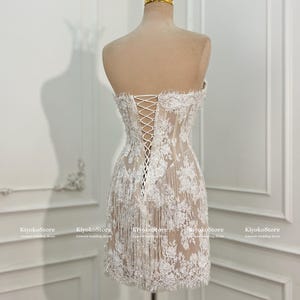 May include: A strapless, short white lace dress featuring a corset-style back with white lacing, intricate floral embroidery, and delicate fringe detailing along the hem. A gold-toned crown sits atop the mannequin displaying the garment.