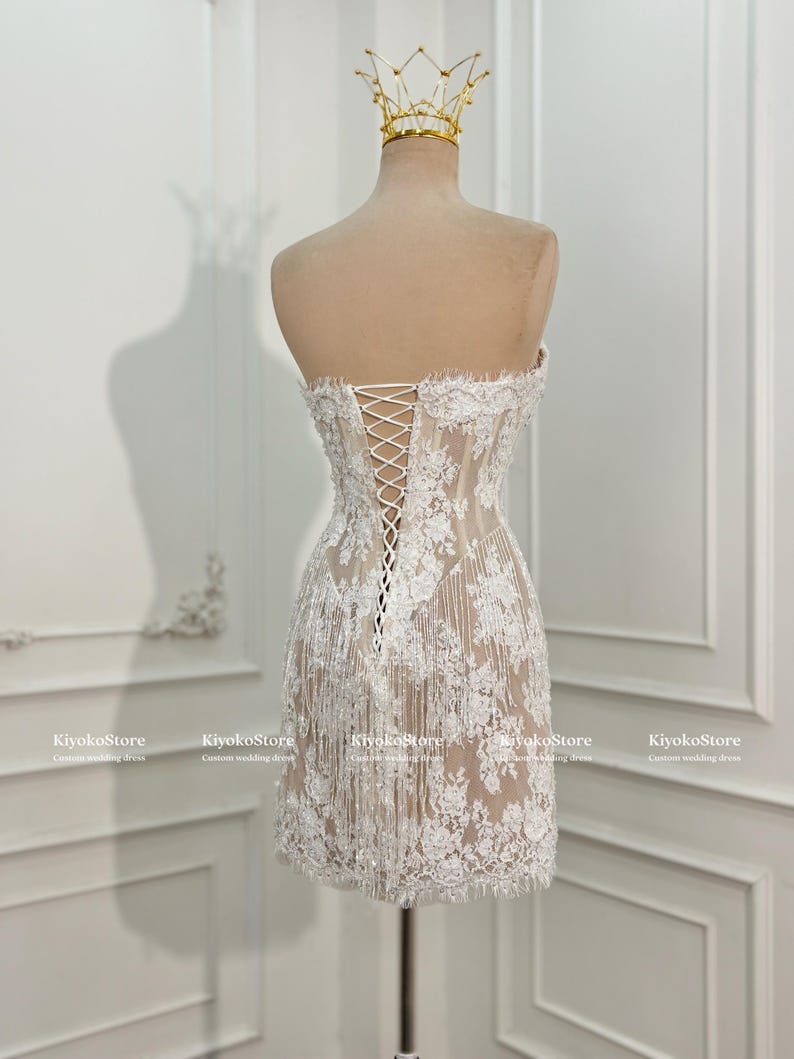 May include: A strapless, short white lace dress featuring a corset-style back with white lacing, intricate floral embroidery, and delicate fringe detailing along the hem. A gold-toned crown sits atop the mannequin displaying the garment.