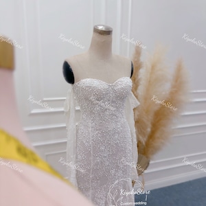 May include: A white strapless wedding dress with a beaded bodice and a flowing skirt. The dress has a fitted silhouette and is adorned with delicate lace details. The dress is displayed on a mannequin against a white background.
