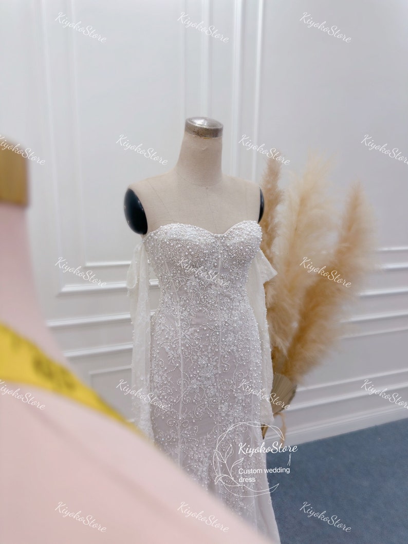May include: A white strapless wedding dress with a beaded bodice and a flowing skirt. The dress has a fitted silhouette and is adorned with delicate lace details. The dress is displayed on a mannequin against a white background.