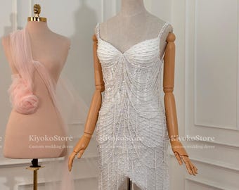 May include: A white, beaded mini dress with spaghetti straps and a V-neckline. The dress is displayed on a mannequin. The dress features intricate beadwork and a layered skirt design. The dress is a custom wedding dress.