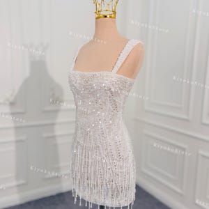 May include: A white, beaded mini dress with a square neckline and spaghetti straps. The dress features intricate beadwork and a fringe hemline. A gold crown sits atop a mannequin head. The dress is approximately 30 inches in length.