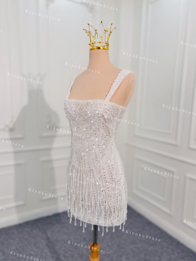 May include: A white, beaded mini dress with a square neckline and spaghetti straps. The dress features intricate beadwork and a fringe hemline. A gold crown sits atop a mannequin head. The dress is approximately 30 inches in length.