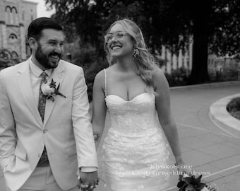 May include: A black and white photo of a smiling couple on their wedding day. The bride wears a short, white, floral-embroidered dress with spaghetti straps. The groom wears a light-colored suit and tie. They are holding hands and walking.