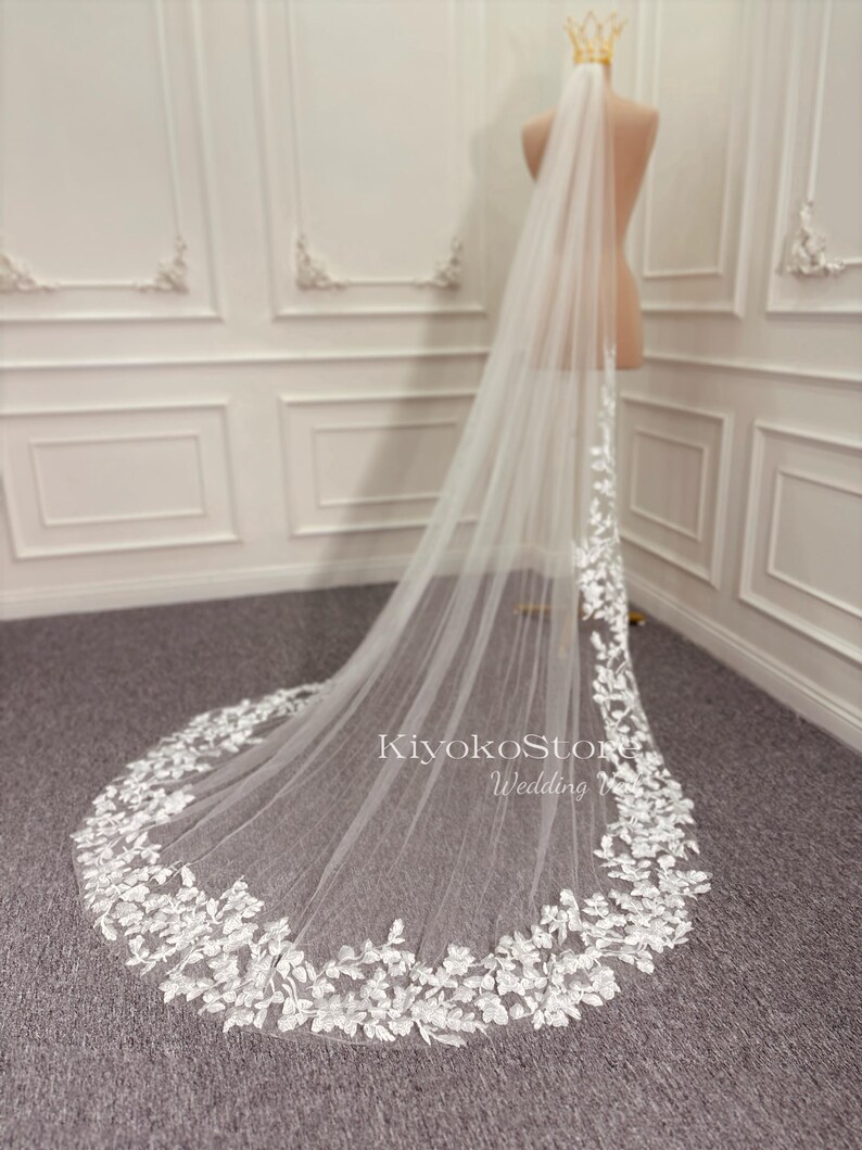 May include: A white bridal veil with a delicate floral lace trim. The veil is long and flowing, with a scalloped edge. The veil is displayed on a mannequin.