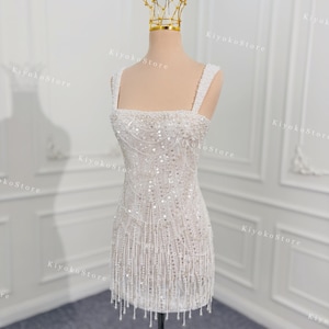 May include: A white, beaded mini dress with a square neckline and fringe detail. The dress is displayed on a mannequin with a gold crown. The dress is embellished with sequins and beads, creating a shimmering effect.